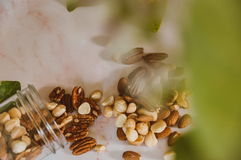 A glass jar of assorted nuts spilled on a marble surface with green leaves in the background.